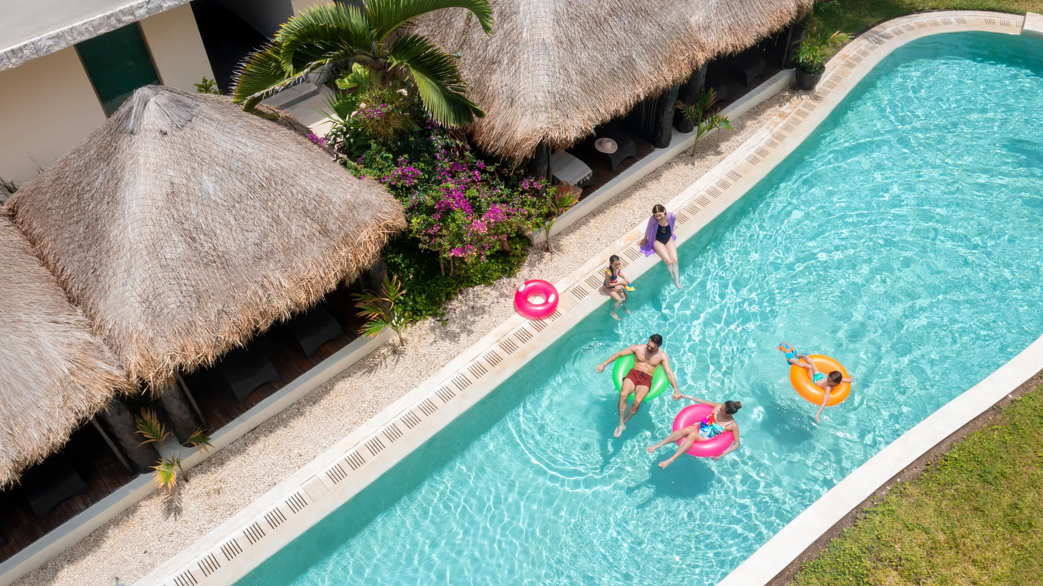 Aerial view of family relaxing and floating in a pool beside thatched-roof cabanas.
