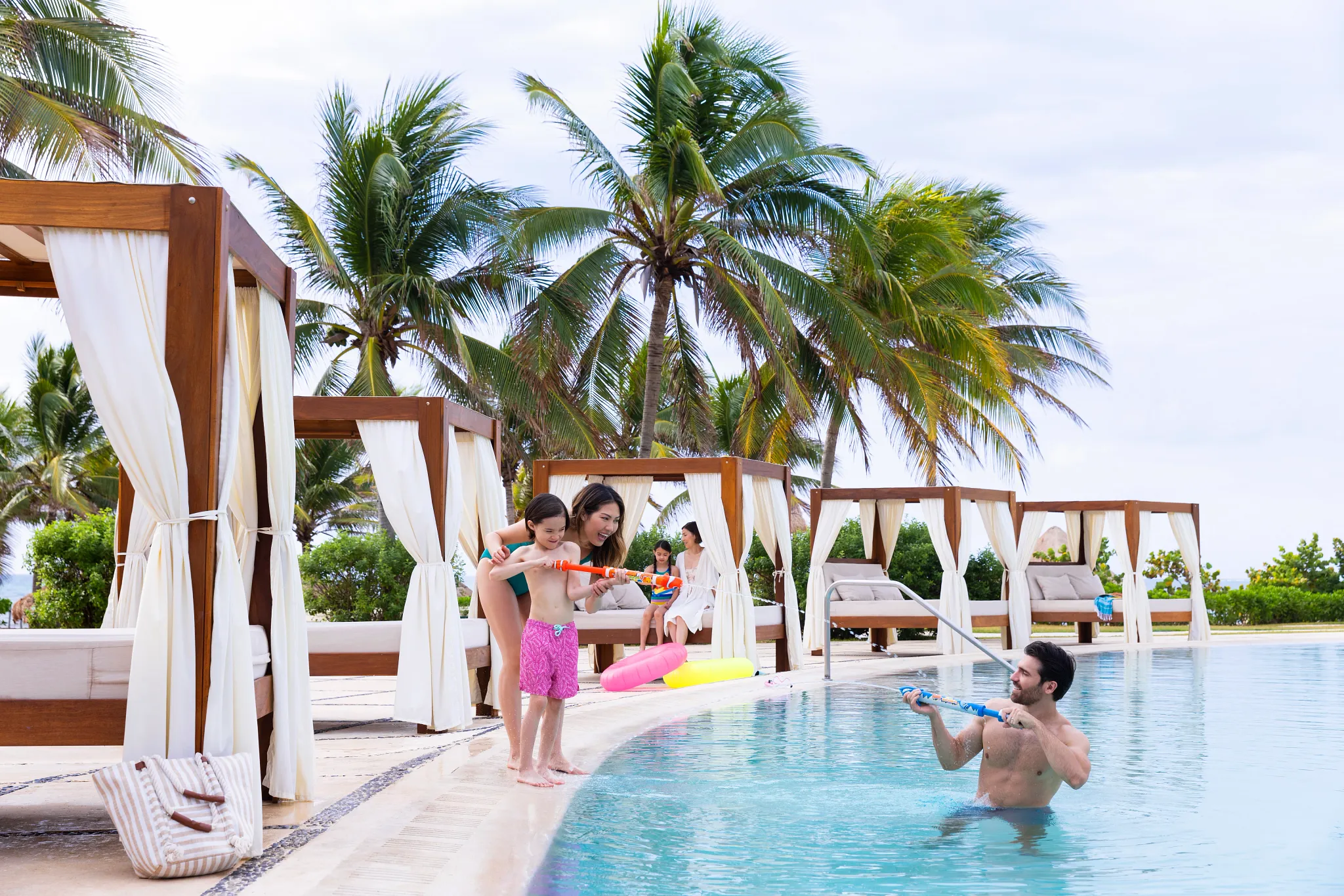 Family playing with water toys in a pool beside beachfront cabanas at Dreams Puerto Morelos Resort & Spa.