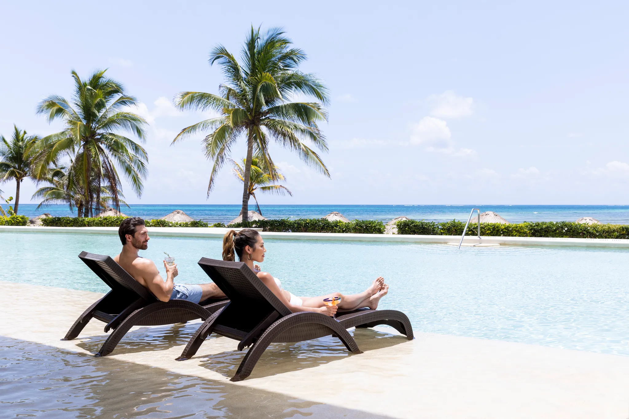 Couple relaxing on in-water loungers with drinks overlooking the ocean at Dreams Puerto Morelos Resort & Spa.