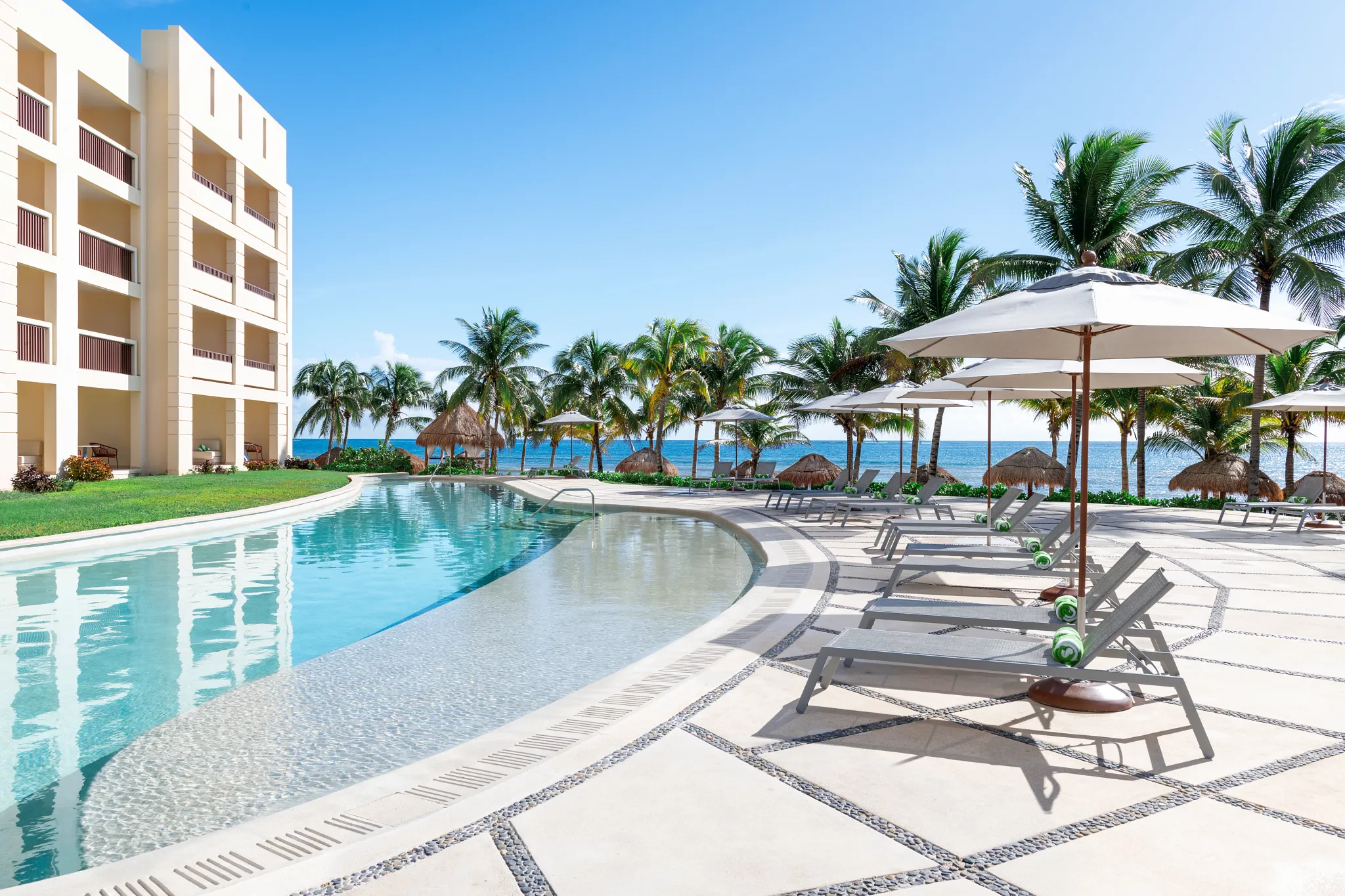 Pool deck with sun loungers and umbrellas next to resort building and ocean views.